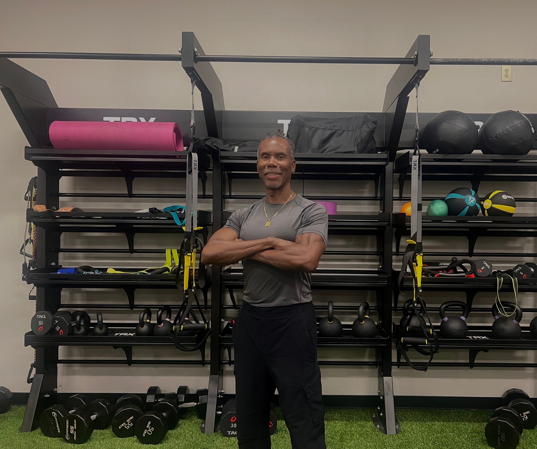 Kenny smiling in the gym, arms crossed, standing in front of a TRX equipment rack